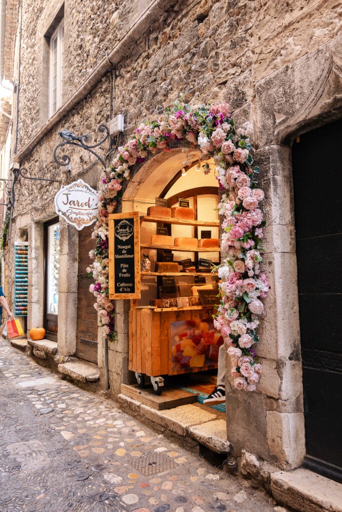 A flower framed shop entrance in Saint Paul de Vence sells sweets and nougat. Signs read "Jarodi Confiserie" "Nougat de Montélimar" "Pâte de fruits" and "Calisson d Aix"