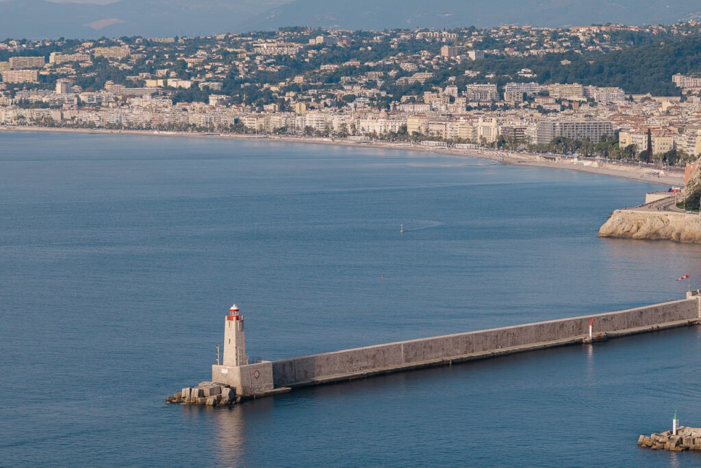 A lighthouse and harbour wall stretch into the blue bay near Nice. The city shoreline curves behind it along the Riviera coast