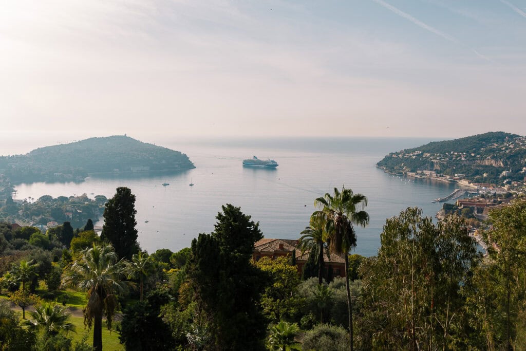 Villefranche sur Mer bay seen from above with a cruise ship anchored between two green headlands. The viewpoint shows the postcard scenery close to Nice.