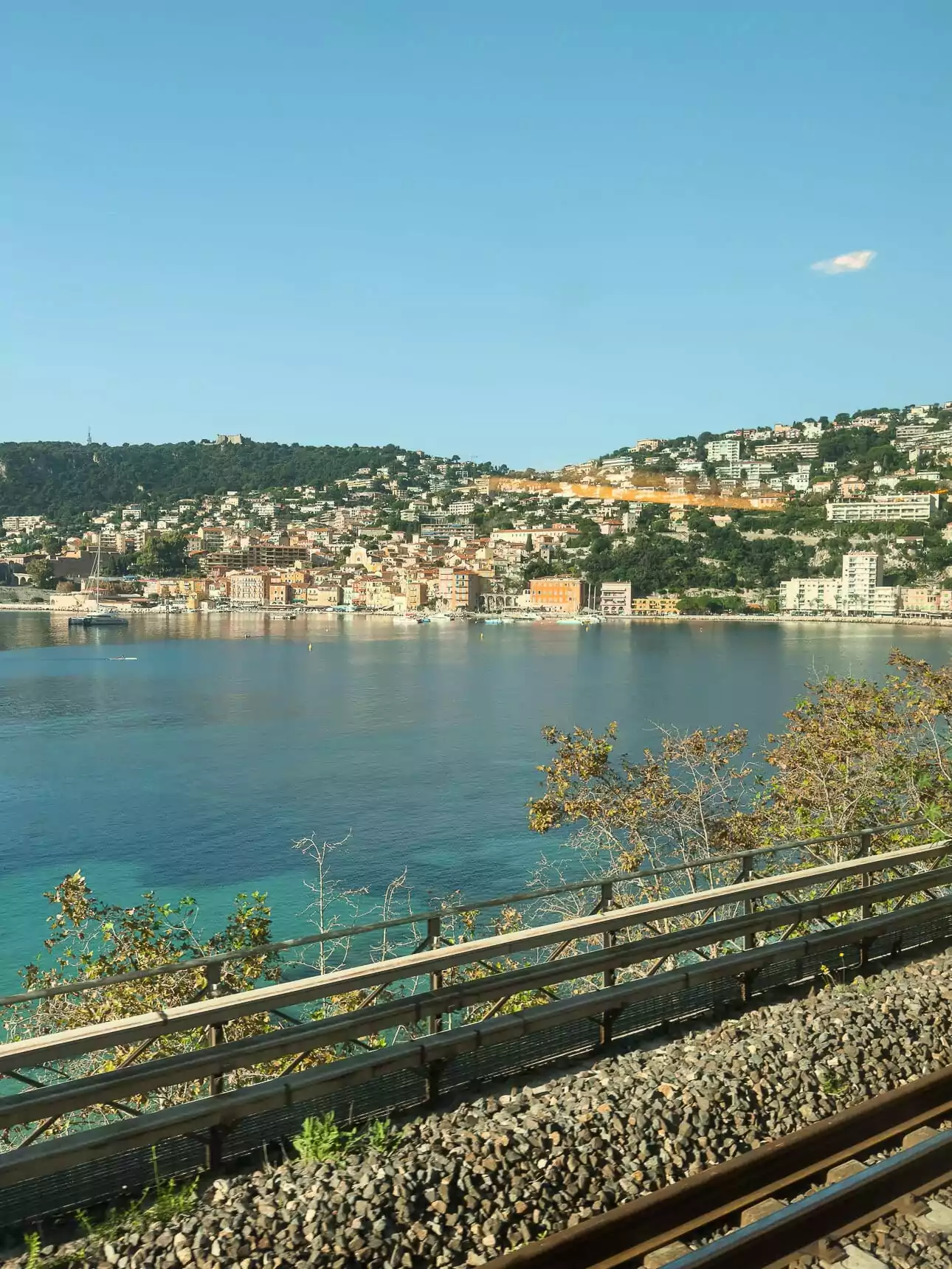 Villefranche sur Mer seen from a train window with blue water and hillside homes beyond the tracks. The view shows why train travel along the French Riviera is so scenic