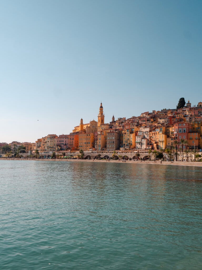 Menton old town rises above the waterfront with pastel buildings and a church tower. The view across the water is a classic stop on a French Riviera by train itinerary