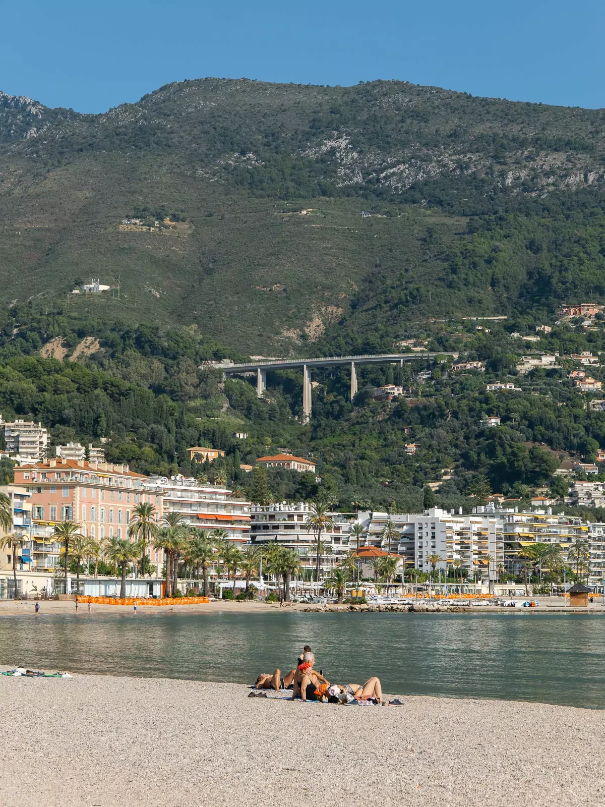 Sunbathers relax on a pebble beach with Menton buildings and green mountains in the background. This coastal scene shows the slower side of travelling the French Riviera by public transport.