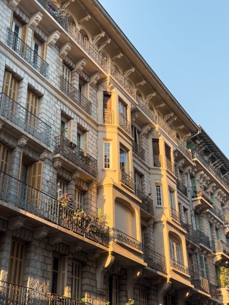 A warm stone building with balconies catches golden light in Nice. The shutters and iron railings give the street a classic Riviera feel