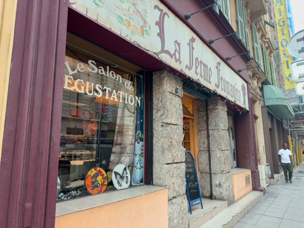 A cheese shop storefront in Nice with burgundy trim and stone columns. Text reads "La Ferme Fromagère" and "Le Salon de Dégustation"