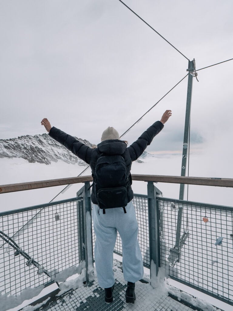 Solo female traveller standing on the outdoor viewing platform at Jungfraujoch with arms raised, looking out over a white snow-covered glacier landscape on an overcast day, dressed in a black puffer jacket and white joggers