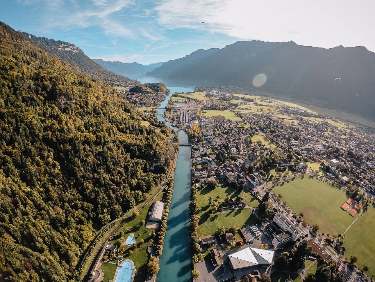 Aerial view of Interlaken, Switzerland, with the turquoise River Aare running through the town centre, surrounded by forested mountains and two lakes visible in the distance, with a paraglider visible in the sky above