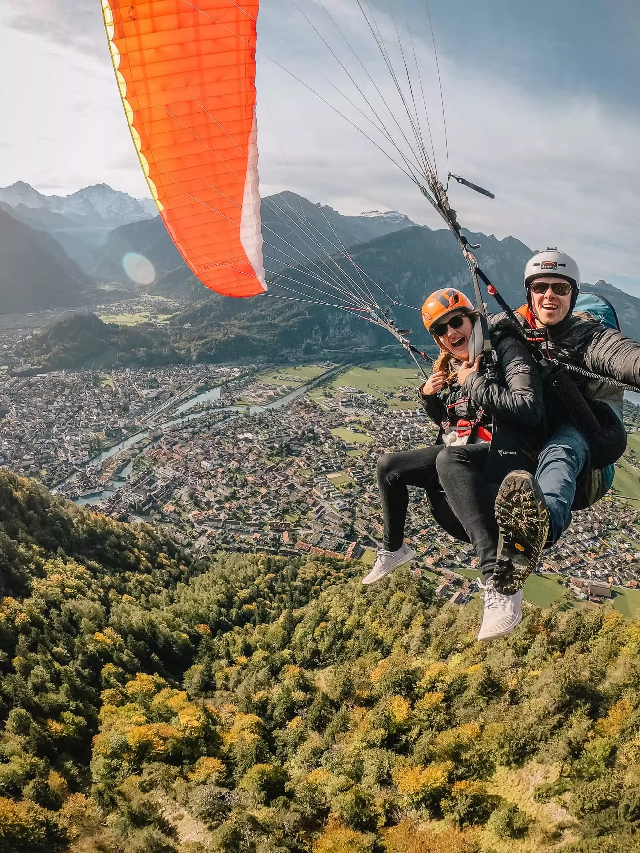 Alexx and a paragliding instructor taking a selfie while flying over Interlaken, with a bright orange parachute above them