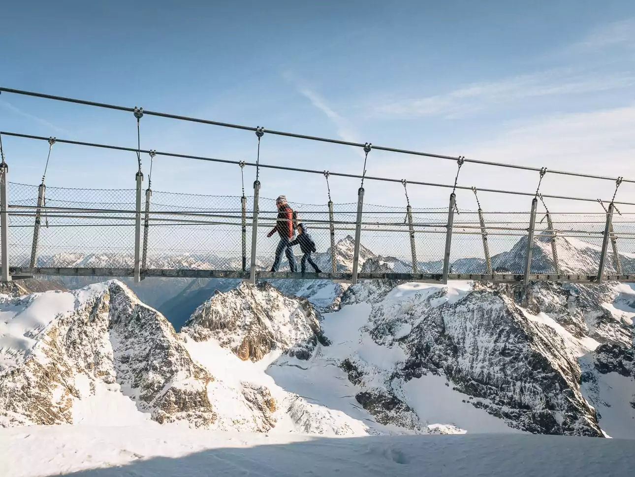 A mother and child cross the Titlis Cliff Walk suspension bridge above snowy mountain peaks