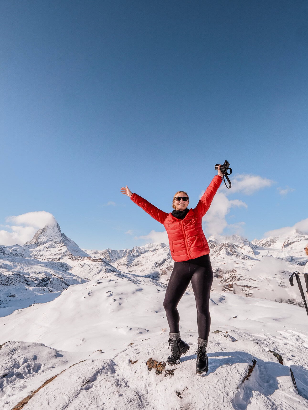 Alexx standing on a snowy rock with her arms up and camera in her hand, with the Matterhorn covered in snow behind her, from the top of Gornergratbahn