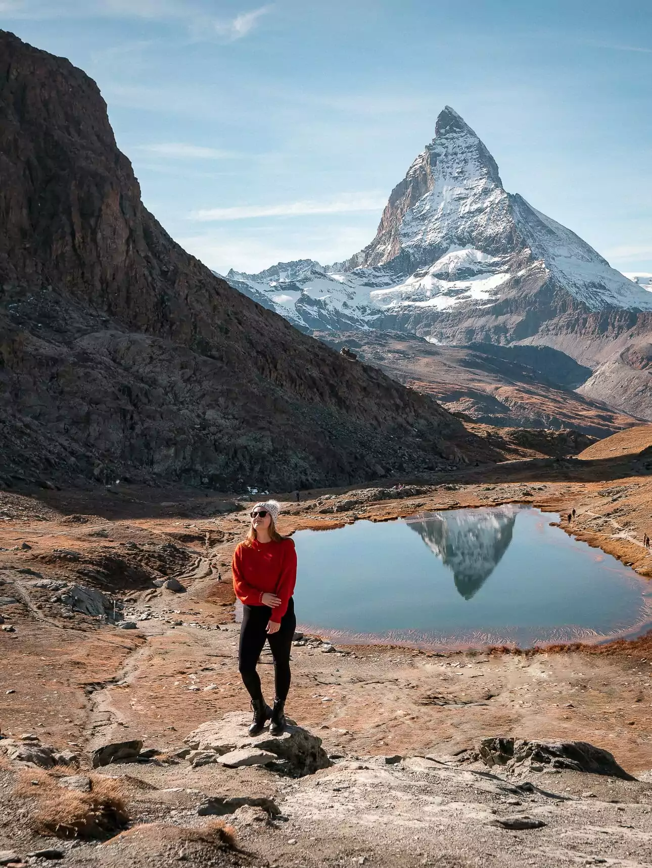 The Matterhorn reflected in a lake near Zermatt, Switzerland, with a solo female traveller standing on a rock in front of it