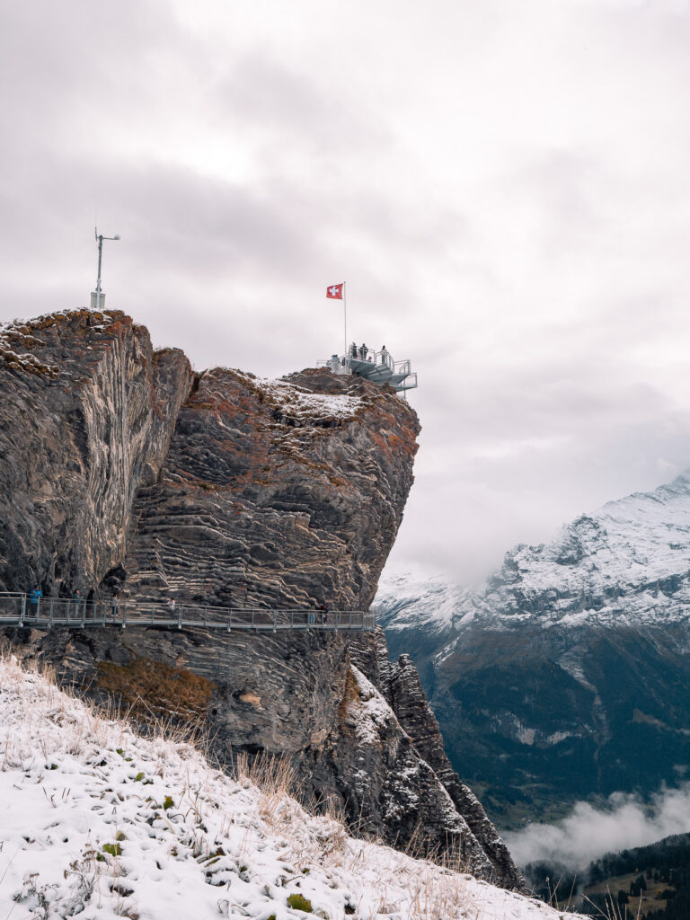 A viewing platform at Grindelwald First jutting out over a steep cliff edge, with a Swiss flag and mountain peaks behind