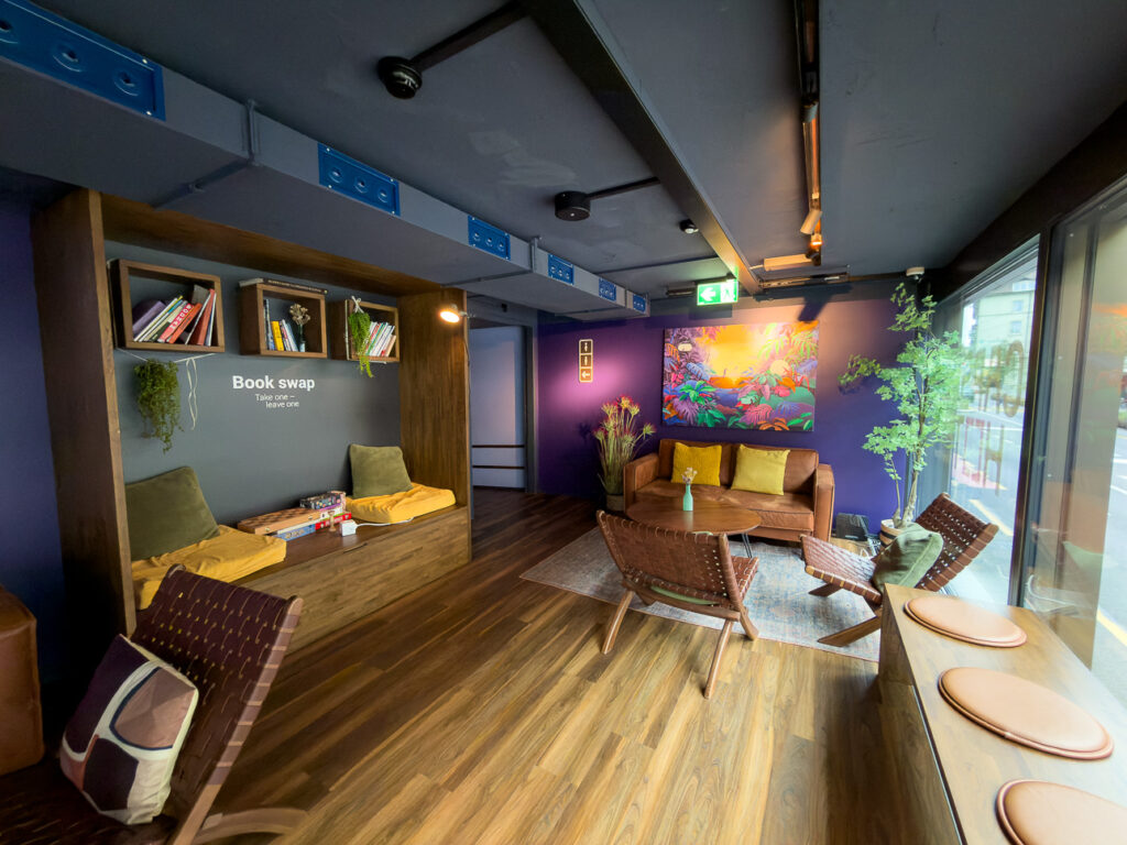 Capsule hotel lounge with a book swap shelf, low seating and a leaf mural on a purple wall