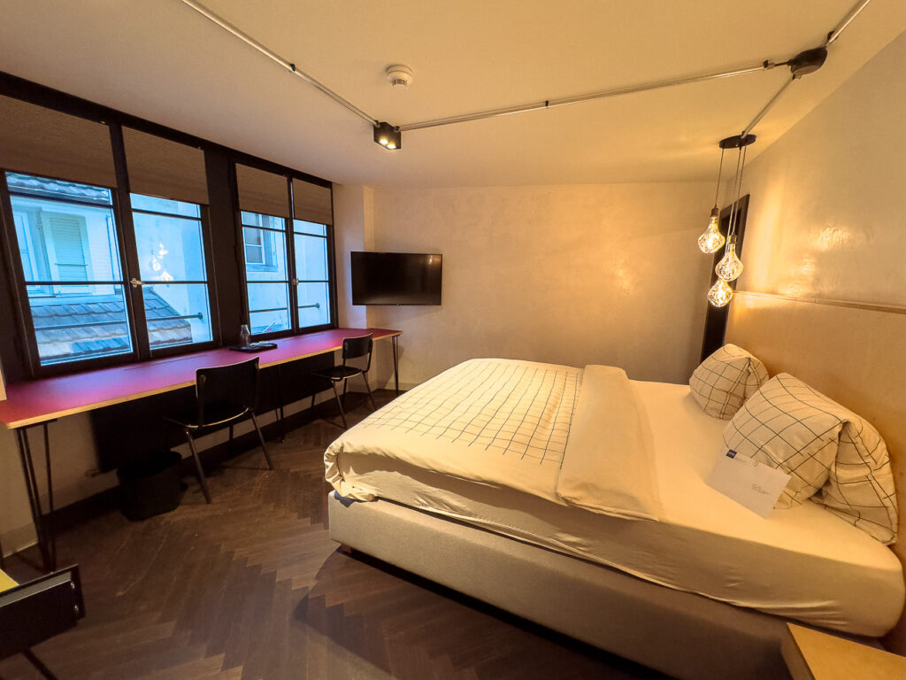 Double room at The Lubo with a bed, wall-mounted TV and a long desk under tall windows