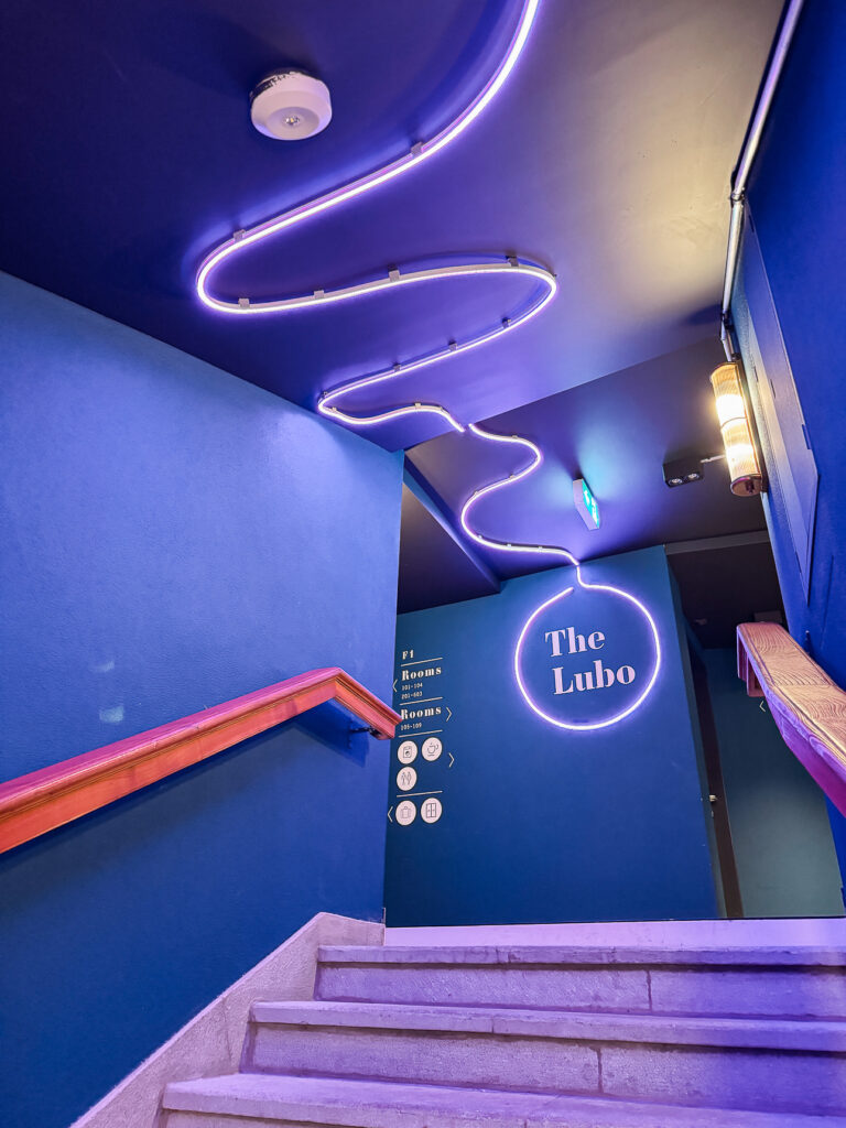 Stairwell at The Lubo with blue walls, room signage and a purple neon strip across the ceiling