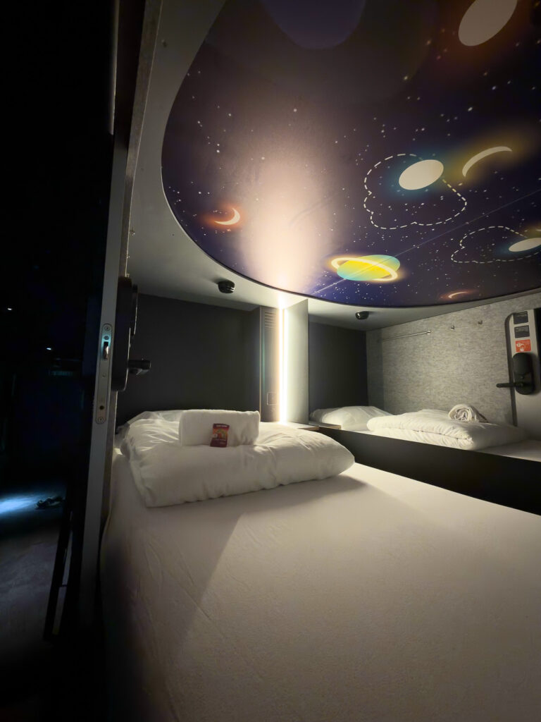 Capsule pod interior with white bedding, a mirrored wall and a planets mural on the ceiling