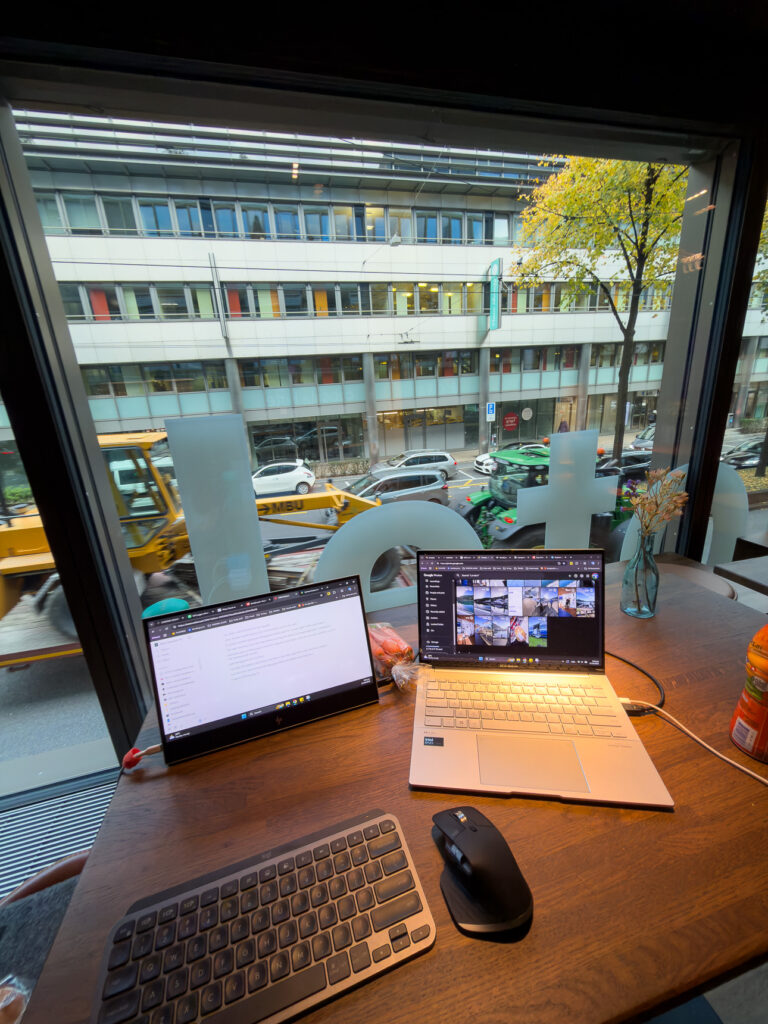 Laptop, portable second screen and keyboard set up on a table beside a window at Capsule Hotel Lucerne Old Town